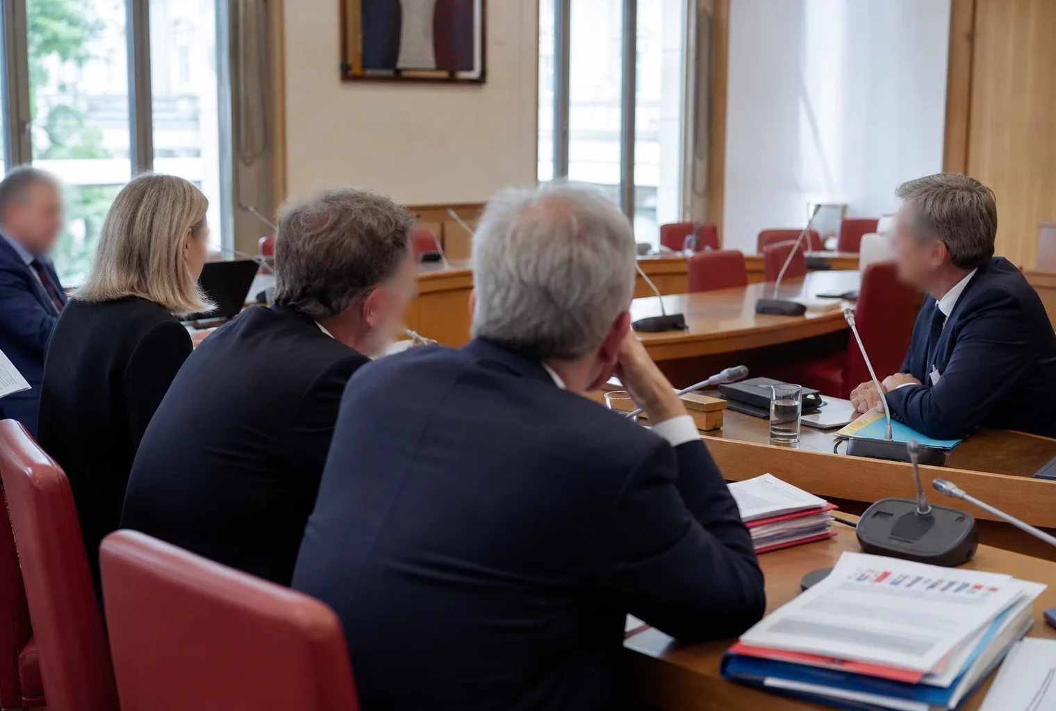 Audition en commission parlementaire avec micros, dossiers et silhouettes anonymes dans une salle claire.