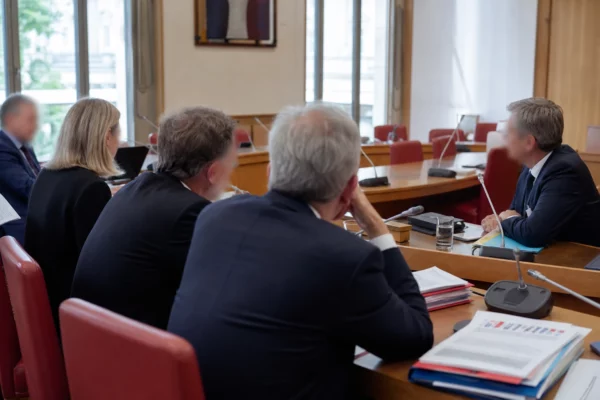 Audition en commission parlementaire avec micros, dossiers et silhouettes anonymes dans une salle claire.