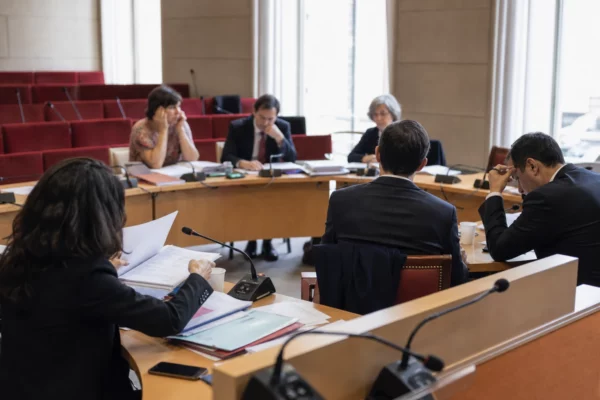 Audition parlementaire en commission avec micros, dossiers ouverts et silhouettes anonymes dans une salle institutionnelle lumineuse.