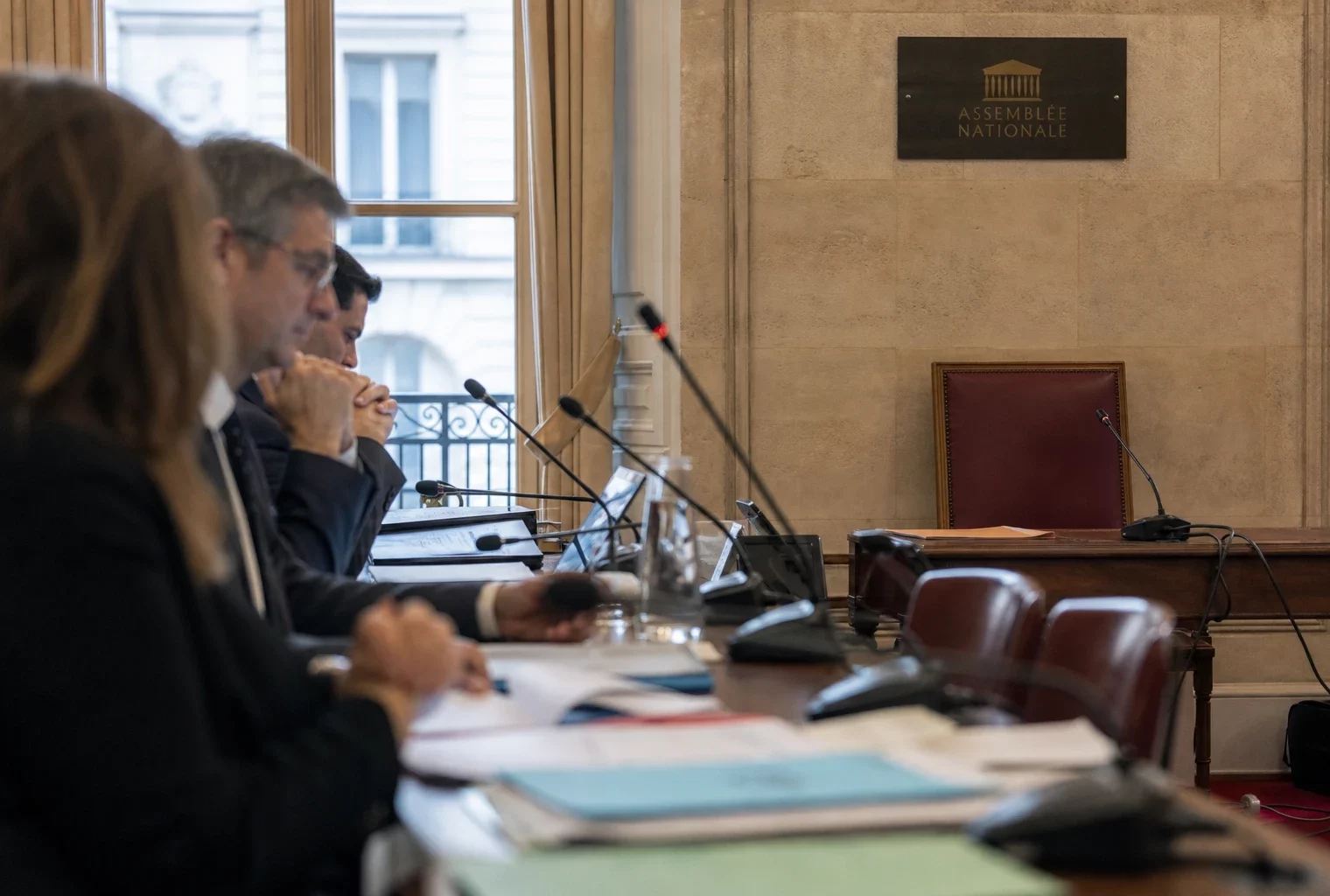 Salle d’audition à l’Assemblée nationale avec micros, dossiers et silhouettes anonymes sous lumière naturelle.