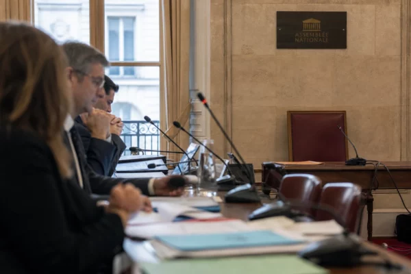 Salle d’audition à l’Assemblée nationale avec micros, dossiers et silhouettes anonymes sous lumière naturelle.