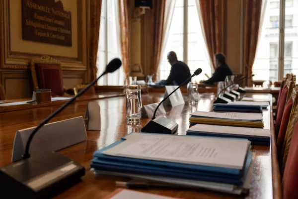 Salle d’audition institutionnelle au Palais Bourbon avec micros, dossiers et silhouettes anonymes sous une lumière claire.