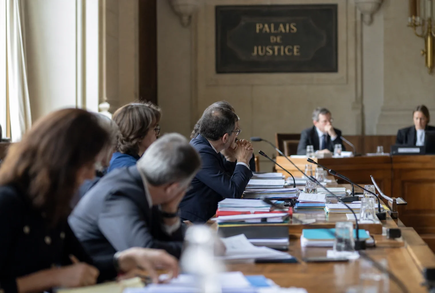 Salle d’audition institutionnelle à Paris avec micros, dossiers ouverts et silhouettes anonymes sous lumière naturelle.