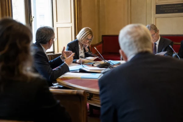 Réunion de commission parlementaire sur l’aide à mourir, avec micros, dossiers et silhouettes anonymes en salle claire.