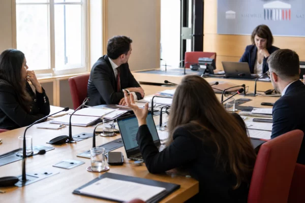 Audition parlementaire en commission avec micros, dossiers et silhouettes anonymes dans une salle claire de l’Assemblée nationale.