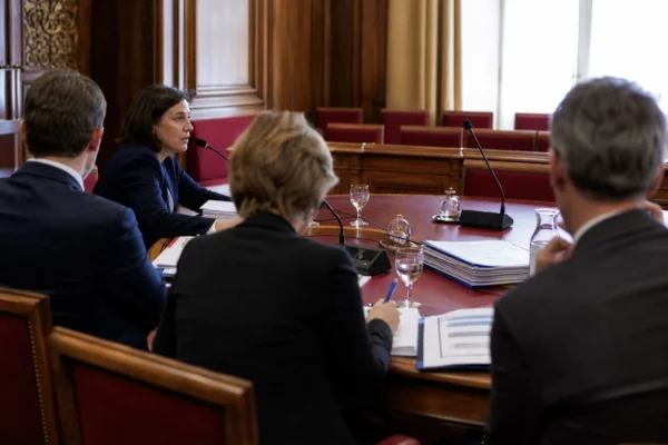 Audition parlementaire en commission avec micros, dossiers ouverts et silhouettes anonymes dans une salle claire
