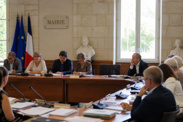 Salle municipale lumineuse avec micros, dossiers et chaise vide, dans une ambiance de conseil local
