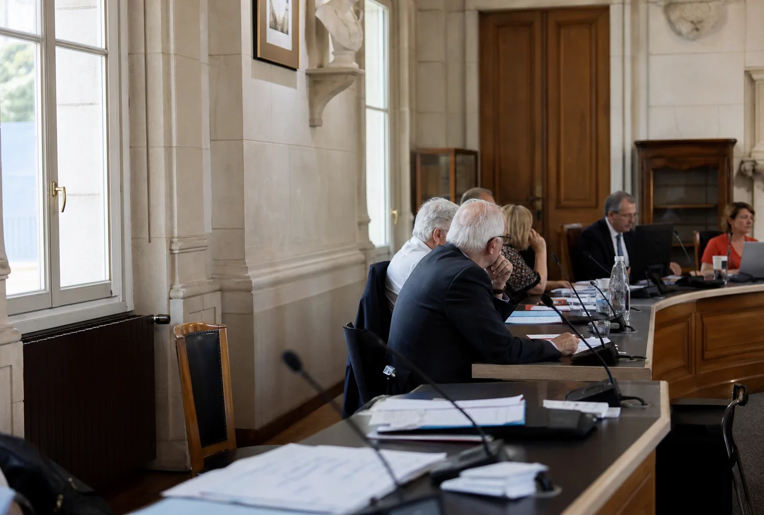 Salle municipale lumineuse avec chaise vide, micros et dossiers flous lors d’une réunion locale sur la canicule.