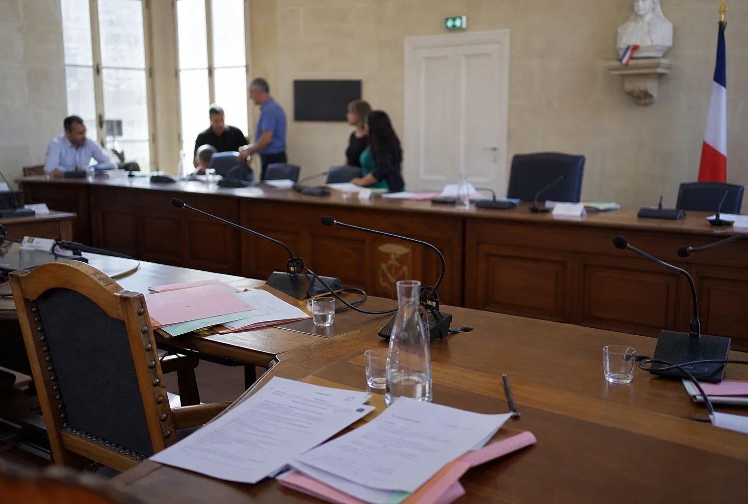 Salle municipale française avec micros, dossiers et chaise vide après une réunion sur la canicule.