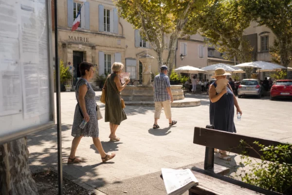Place de village du sud de la France sous forte chaleur, habitants anonymes près d’une mairie et d’une fontaine