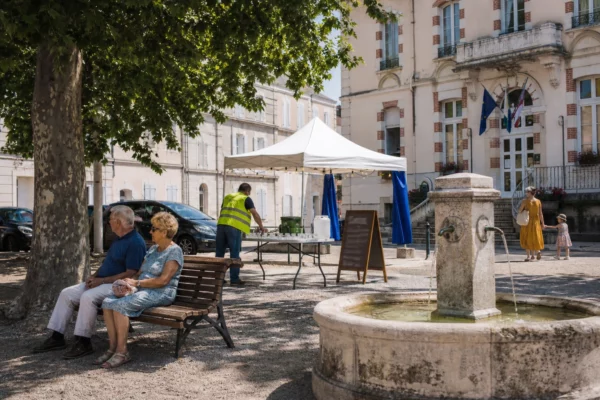 Place de ville moyenne en pleine canicule, habitants anonymes à l’ombre près d’une mairie et d’un point d’eau.