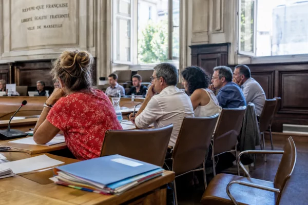 Salle municipale lumineuse avec micros, dossiers flous et chaise vide pendant une réunion sur la canicule
