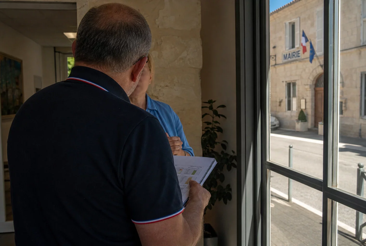 Un élu local de dos consulte un carnet devant une mairie, dans une ambiance de canicule et de débat politique.