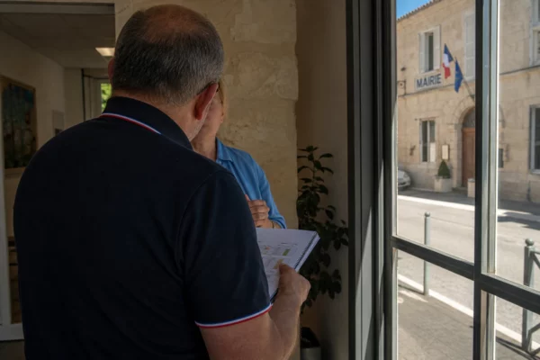 Un élu local de dos consulte un carnet devant une mairie, dans une ambiance de canicule et de débat politique.