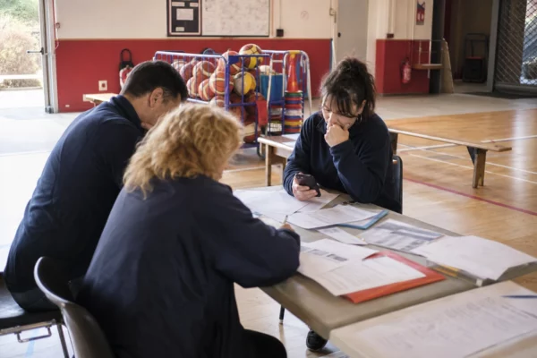 Dans un gymnase municipal, des membres d’un club local consultent des papiers autour d’une table, en lumière naturelle.