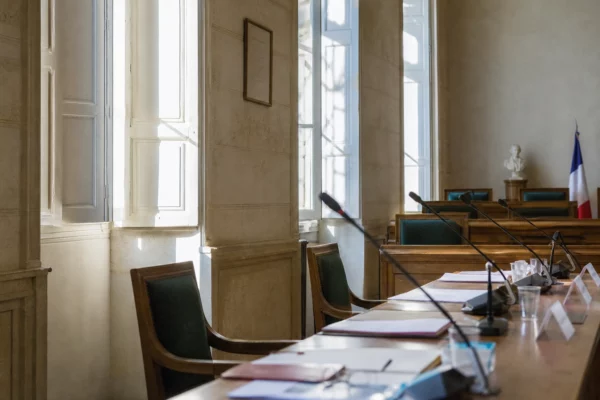 Salle municipale claire avec chaise vide, micros et dossiers flous, ambiance de réunion perturbée par la canicule.