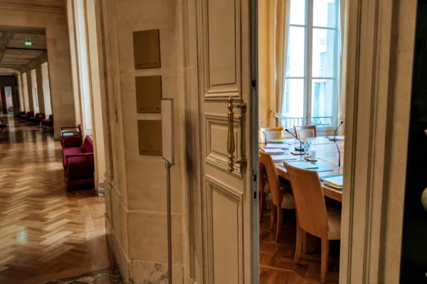 Couloir du Palais Bourbon avec salle de commission vide, micros et dossiers, lumière naturelle claire.