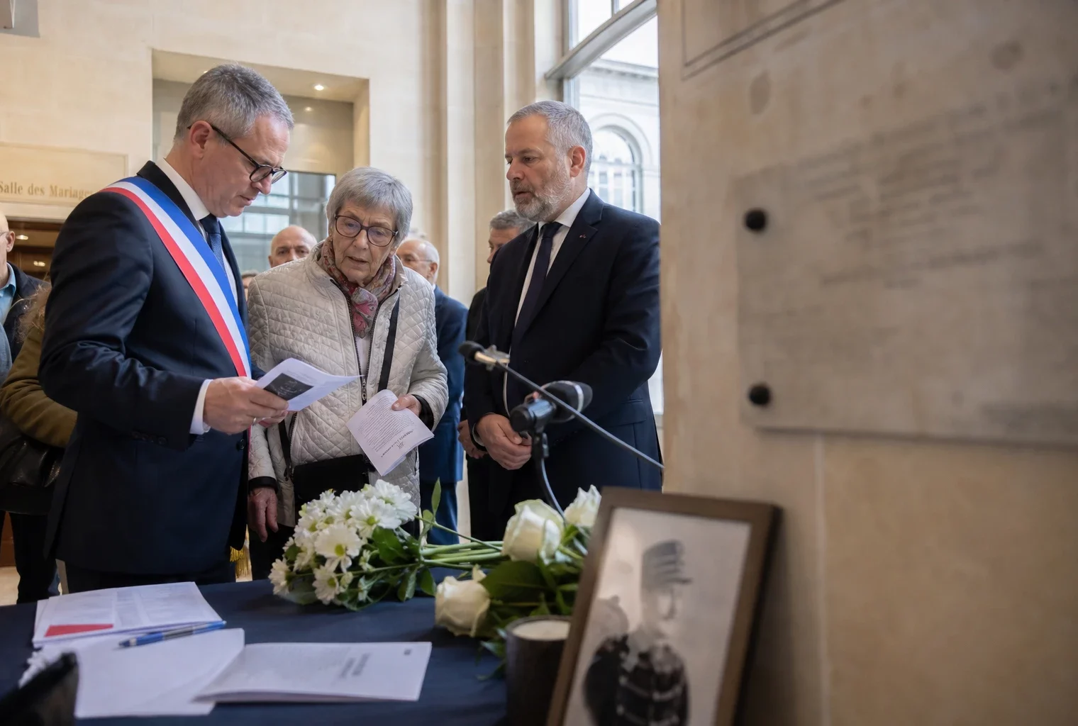 Cérémonie commémorative à Dreyfus dans une salle civique française avec plusieurs participants anonymes et fleurs blanches