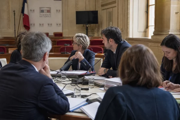 Réunion de commission à l’Assemblée nationale avec micros, dossiers ouverts et silhouettes anonymes en discussion.
