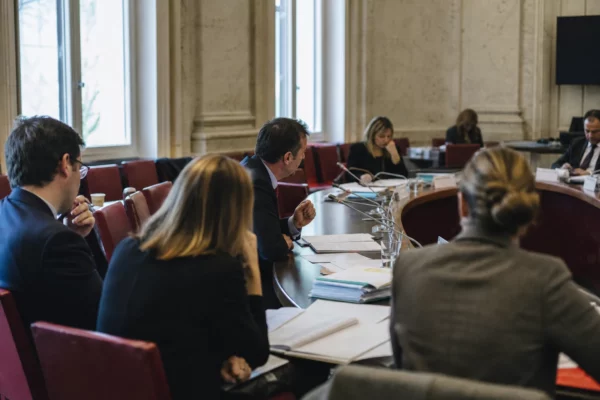Réunion de commission parlementaire avec micros, dossiers ouverts et élus anonymes dans une salle lumineuse.