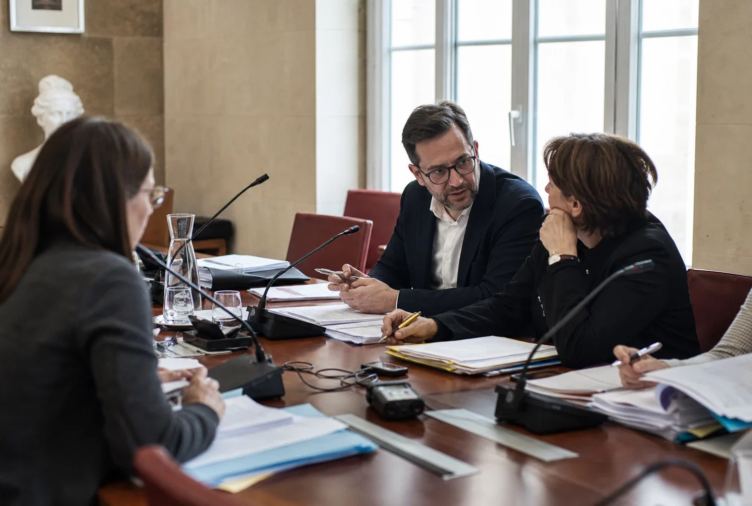 Réunion de commission municipale en France avec micros, dossiers ouverts et silhouettes anonymes autour d’une table.