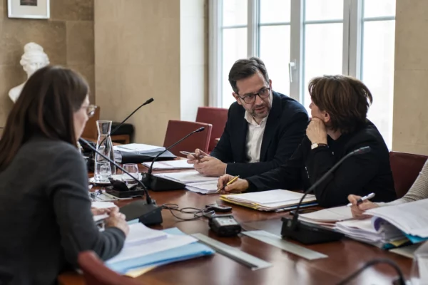 Réunion de commission municipale en France avec micros, dossiers ouverts et silhouettes anonymes autour d’une table.
