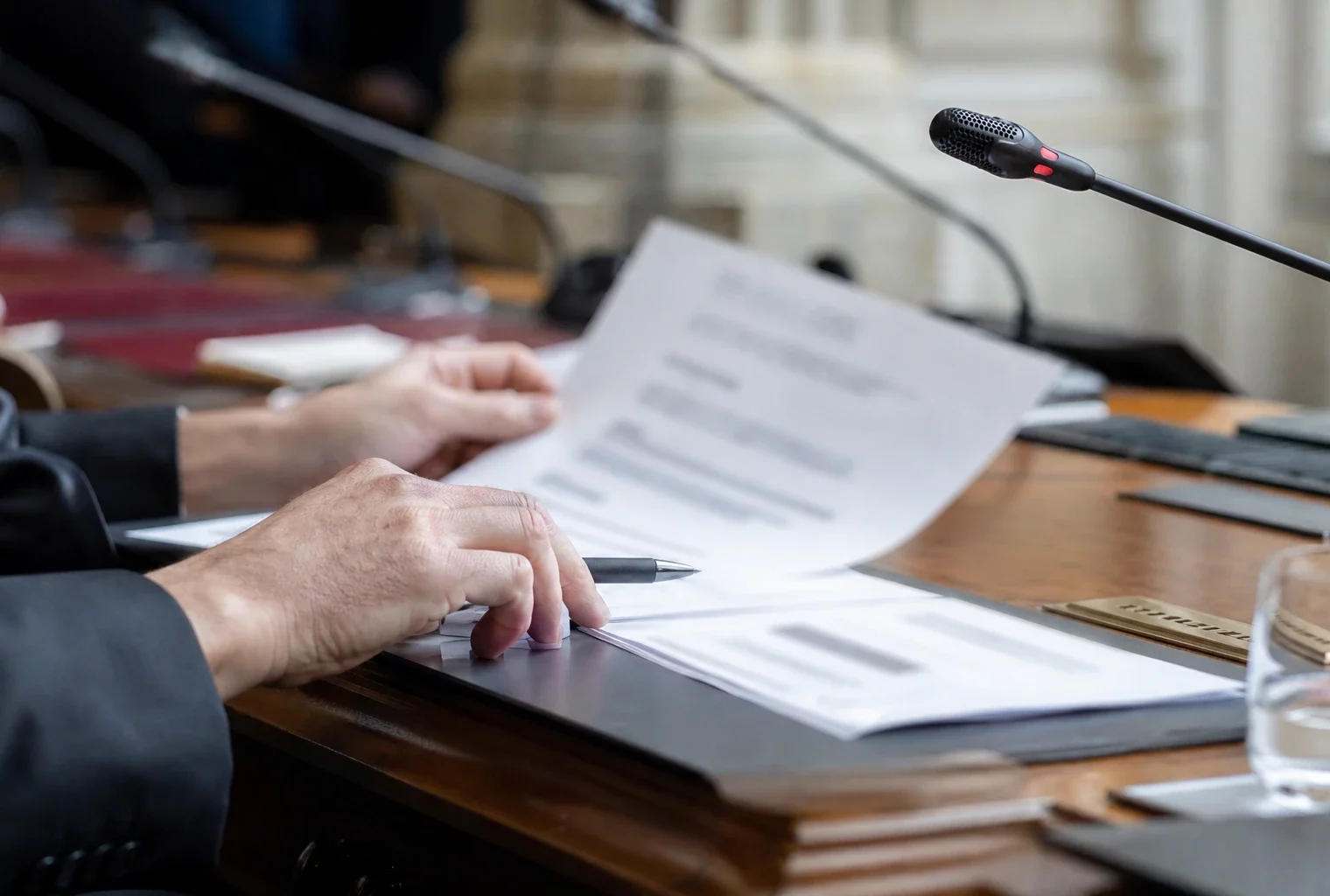Dossier flou et micro de commission sur une table de hearing parlementaire, mains anonymes en lumière naturelle.
