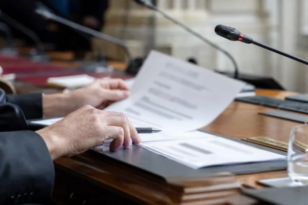 Dossier flou et micro de commission sur une table de hearing parlementaire, mains anonymes en lumière naturelle.