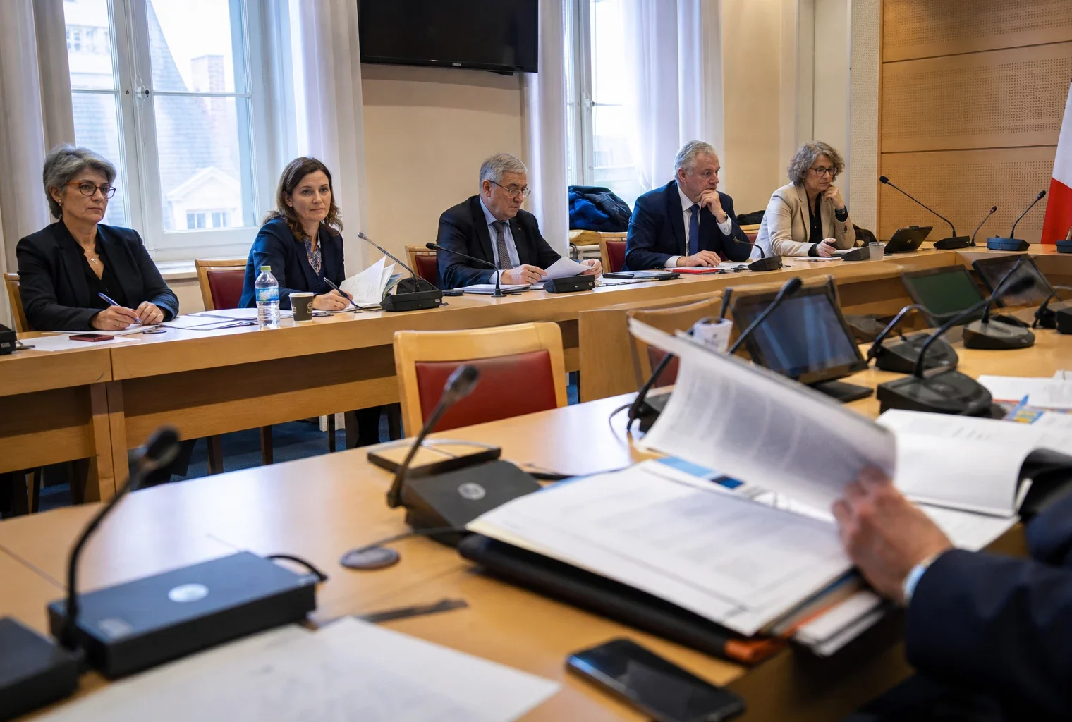 Réunion de commission parlementaire avec micros, dossiers ouverts et silhouettes anonymes dans une salle lumineuse.