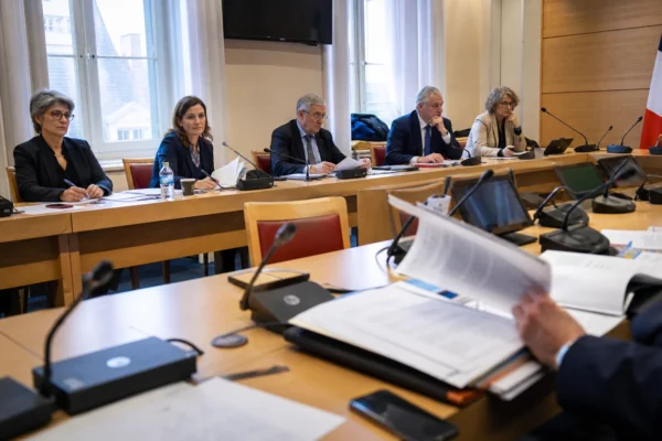 Réunion de commission parlementaire avec micros, dossiers ouverts et silhouettes anonymes dans une salle lumineuse.