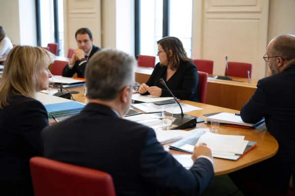 Réunion de commission parlementaire en France avec micros, dossiers et silhouettes anonymes autour d’une table.