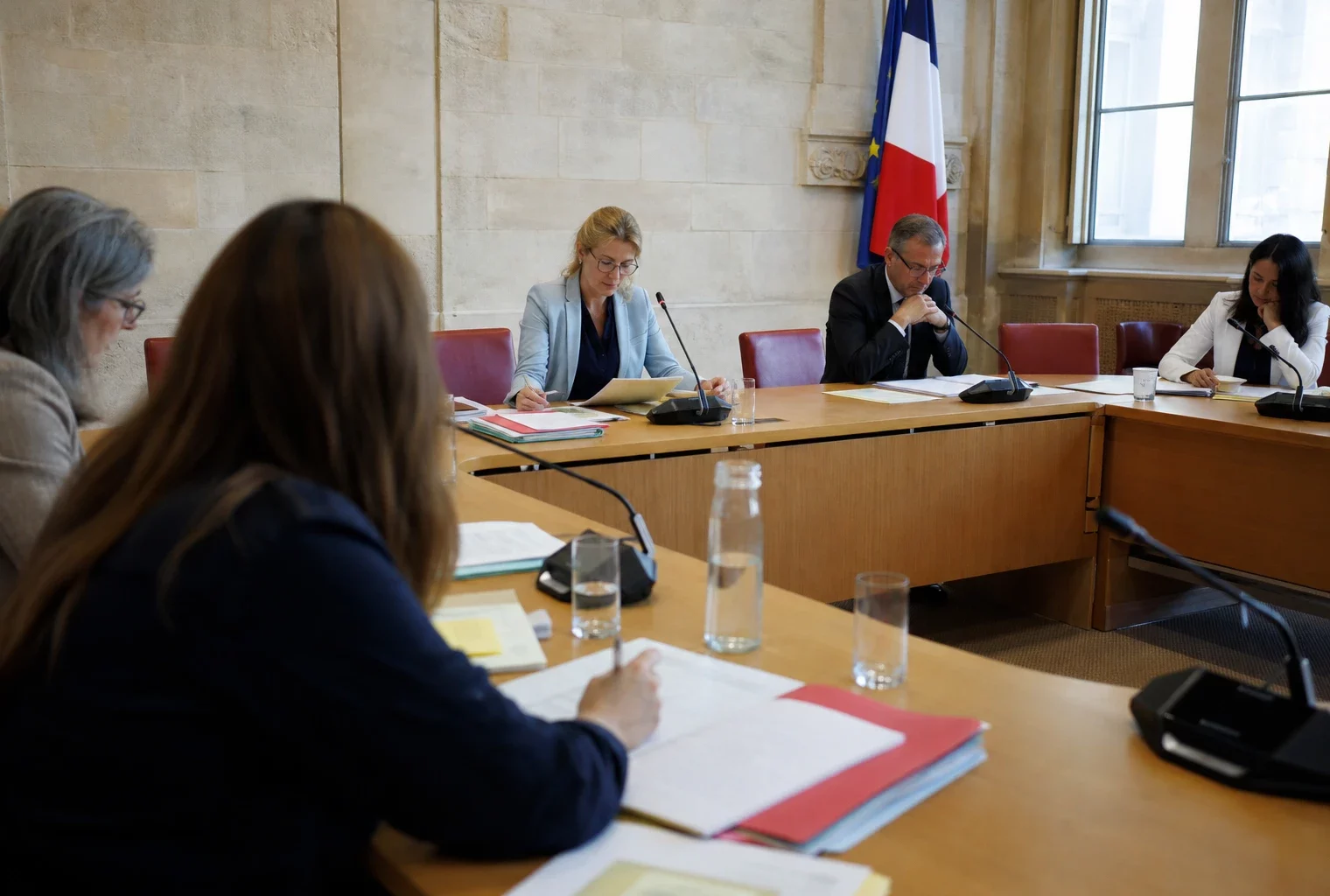 Réunion de commission parlementaire avec micros, dossiers ouverts et silhouettes anonymes dans une salle lumineuse.