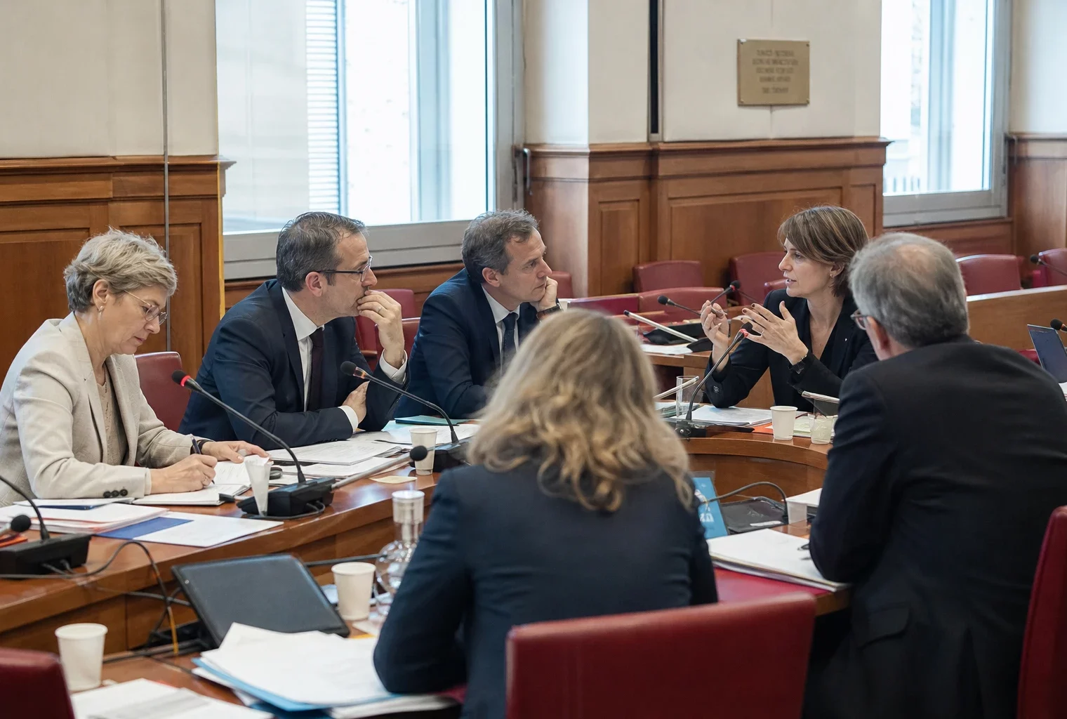 Réunion parlementaire dans une commission, avec micros, dossiers ouverts et plusieurs personnes anonymes en discussion