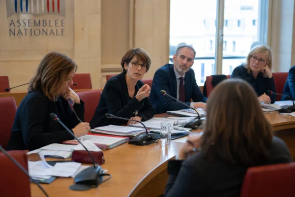 Réunion de commission parlementaire avec micros, dossiers ouverts et silhouettes anonymes sous une lumière claire.
