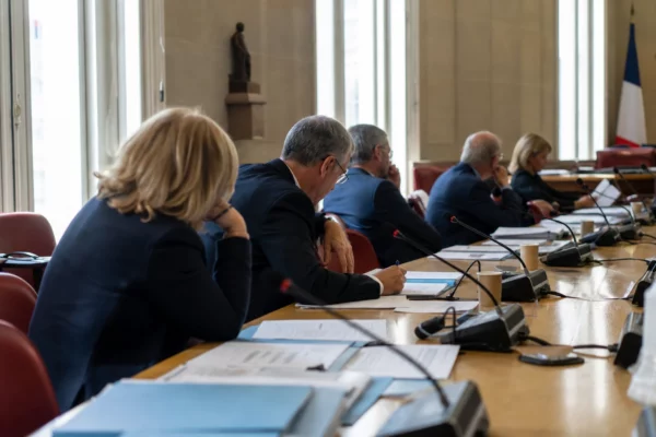 Réunion parlementaire française en commission, avec micros, dossiers et silhouettes anonymes dans une salle lumineuse.
