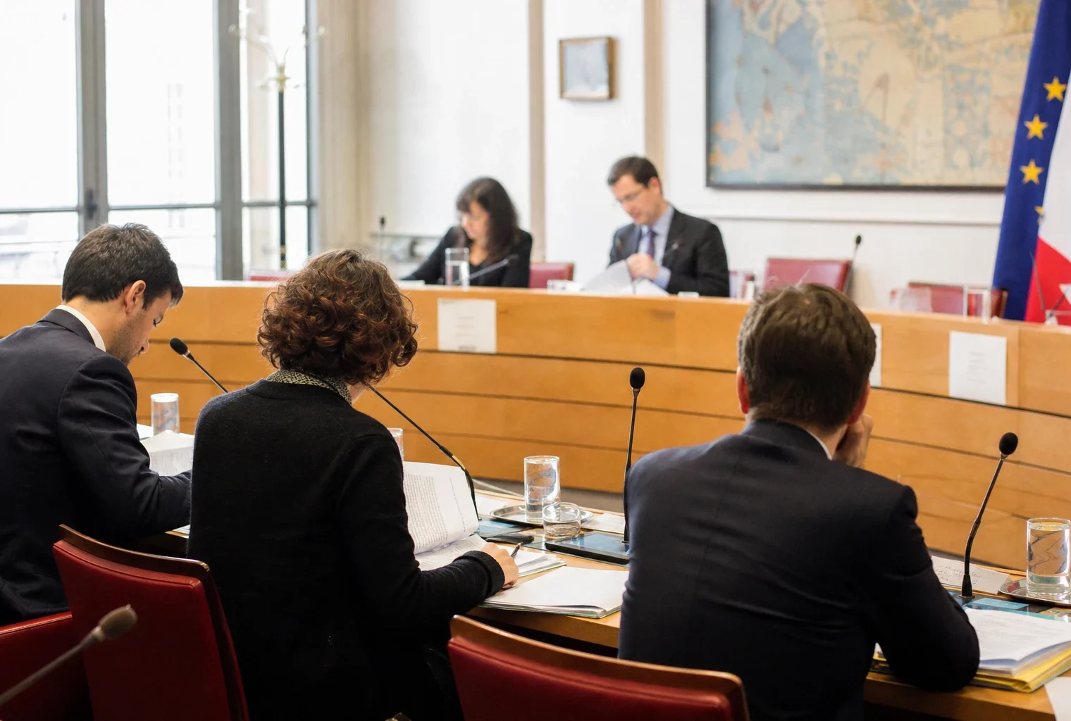 Réunion de commission parlementaire avec micros, dossiers ouverts et silhouettes anonymes dans une salle lumineuse