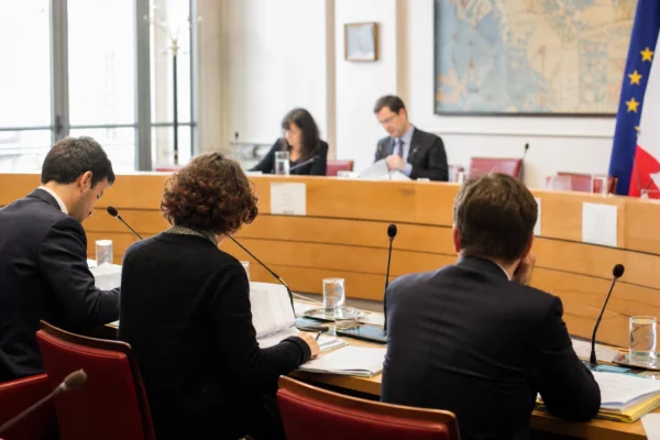 Réunion de commission parlementaire avec micros, dossiers ouverts et silhouettes anonymes dans une salle lumineuse