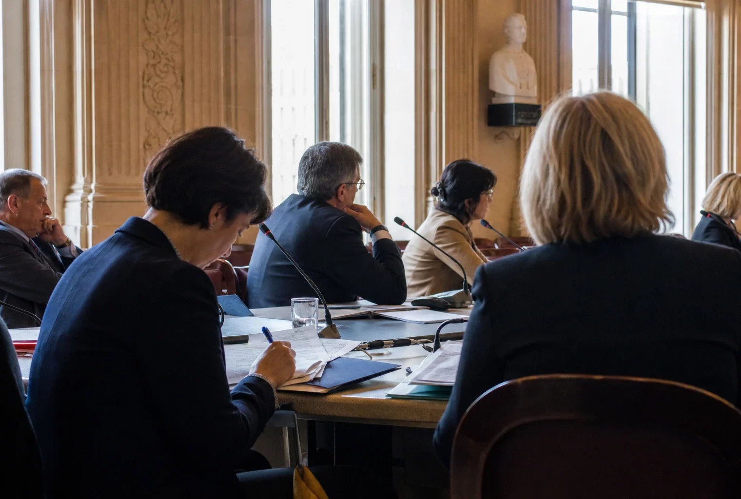 Réunion de commission parlementaire avec micros, dossiers ouverts et silhouettes anonymes autour d’une table
