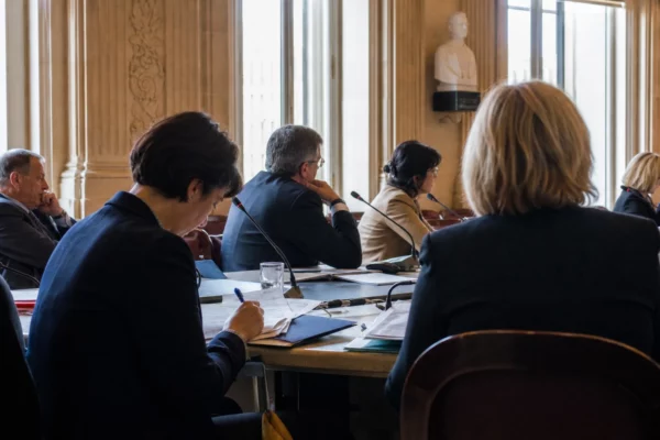 Réunion de commission parlementaire avec micros, dossiers ouverts et silhouettes anonymes autour d’une table