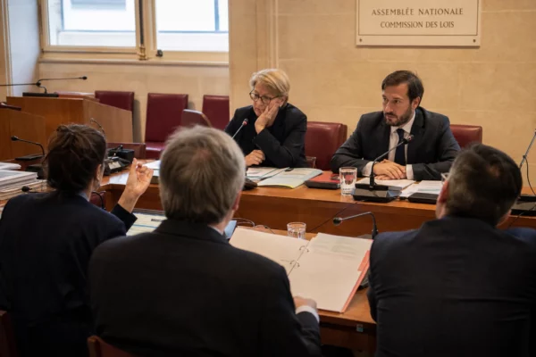 Réunion de commission parlementaire avec micros, dossiers et silhouettes anonymes dans une salle institutionnelle lumineuse.