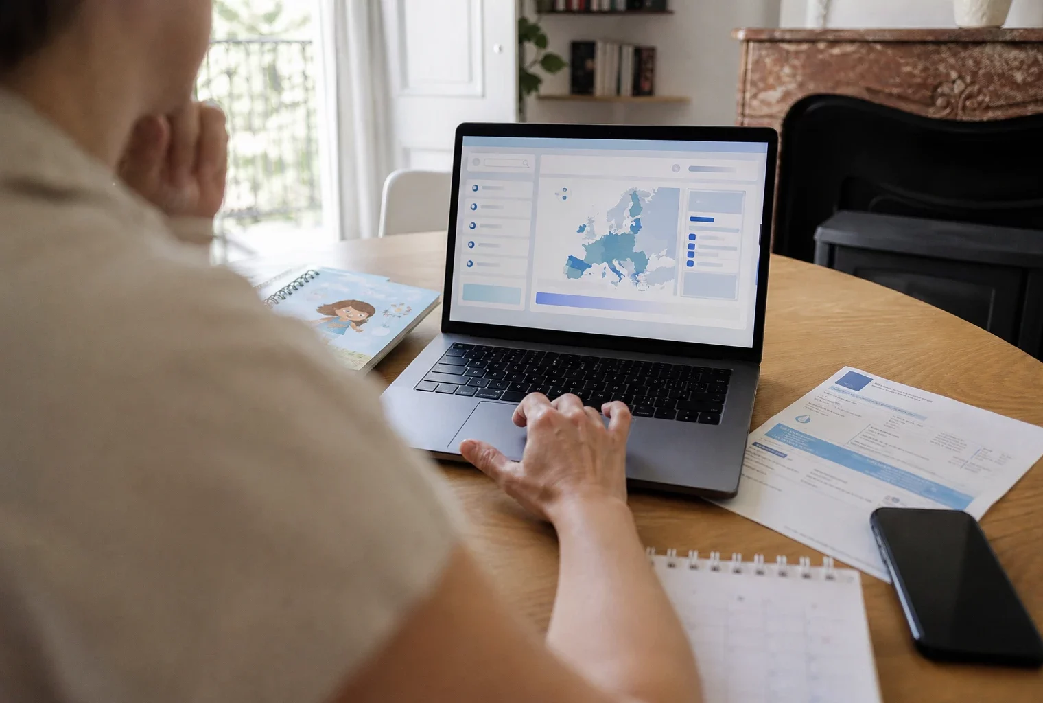 Vue épaule sur un ordinateur affichant une interface abstraite, avec main floue et documents de foyer sur un bureau clair.
