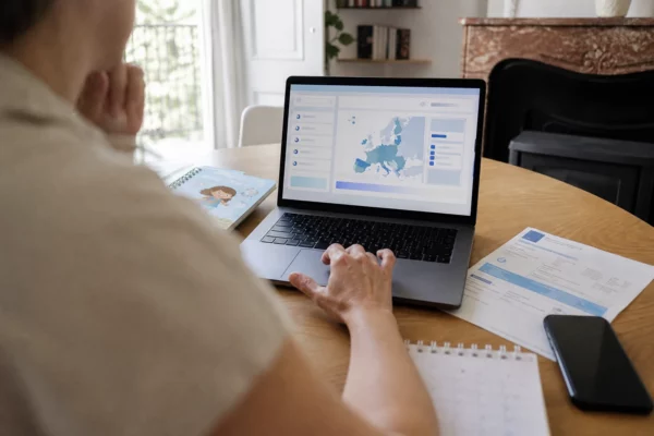 Vue épaule sur un ordinateur affichant une interface abstraite, avec main floue et documents de foyer sur un bureau clair.