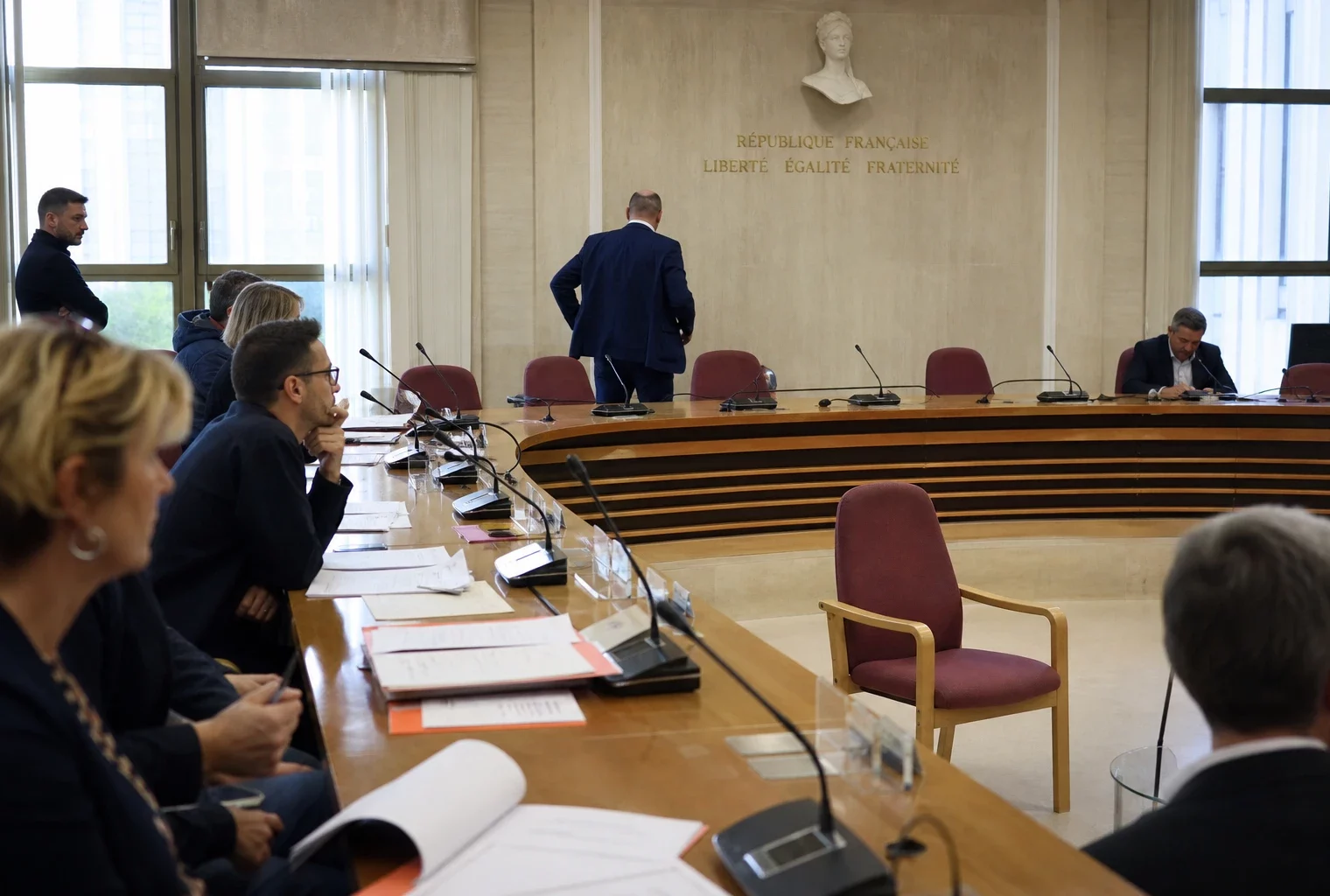 Salle de conseil municipal en France avec chaise vide, micros et dossiers flous sous une lumière naturelle claire.