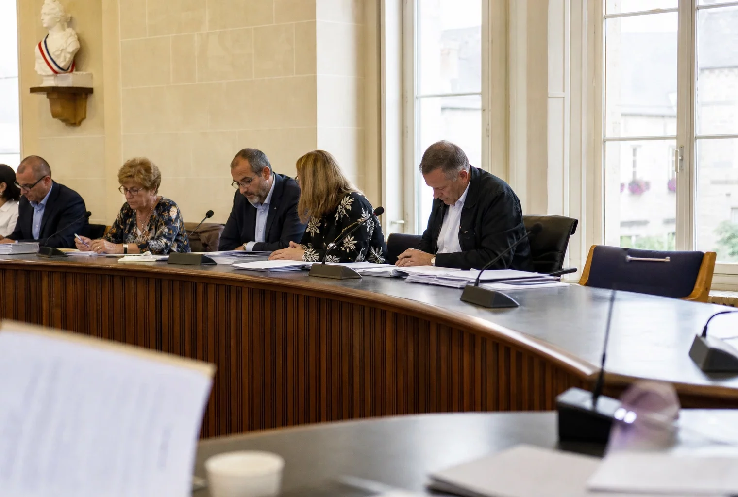 Salle municipale vue en oblique avec chaise vide, micros de table et dossiers flous, ambiance institutionnelle calme.