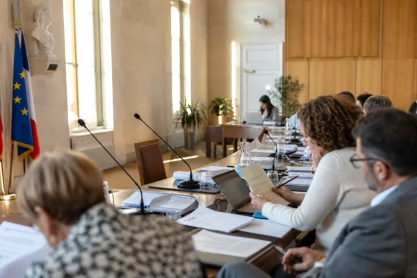 Salle municipale lumineuse avec micros, dossiers flous et chaise vide, dans une ambiance de conseil local.
