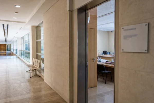 Couloir lumineux d’un bâtiment institutionnel avec une salle d’audition visible au fond, ambiance de reportage politique.
