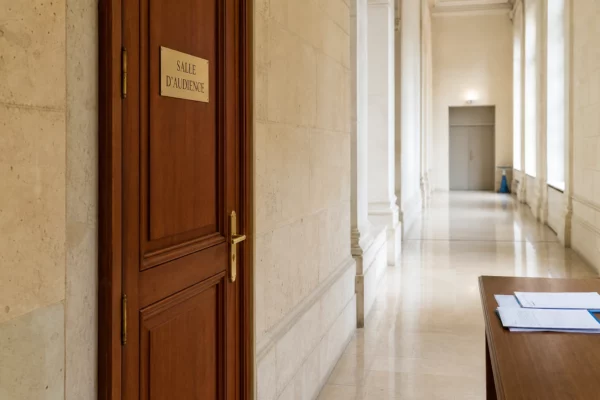 Couloir institutionnel lumineux menant à une salle d’audition, dans un décor officiel français sobre et vide.