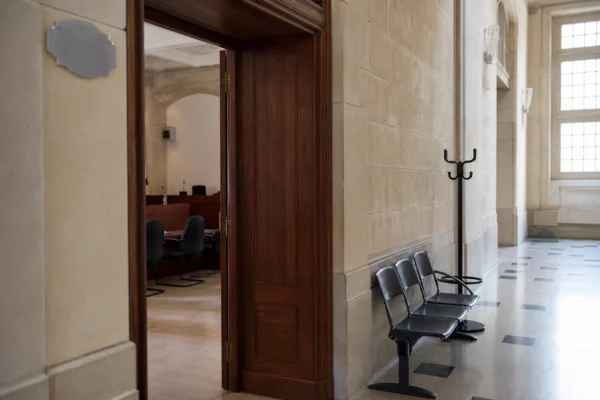 Couloir clair d’un bâtiment institutionnel français menant à une salle d’audition, ambiance sobre et documentaire.
