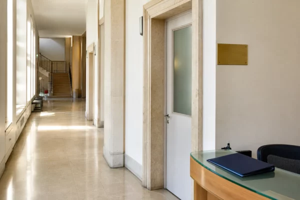 Couloir institutionnel clair avec porte sobre et hall officiel vide, en lumière naturelle, style photojournalistique.