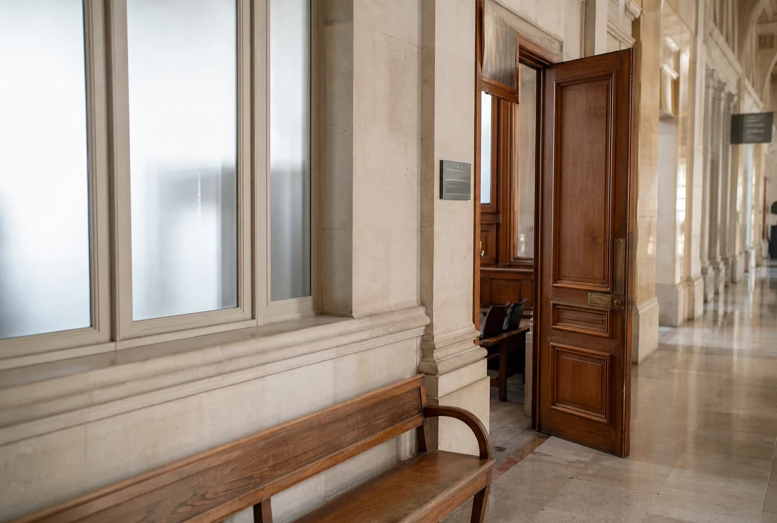 Couloir clair d’un tribunal parisien avec porte d’audience entrouverte et lumière naturelle sur les murs de pierre.